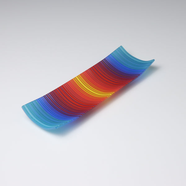 A decorative shallow rectangular boat shaped glass plate with raised corners and a  colourful ColourWave stripe patterns showing key colours of cool blues warm reds and yelows