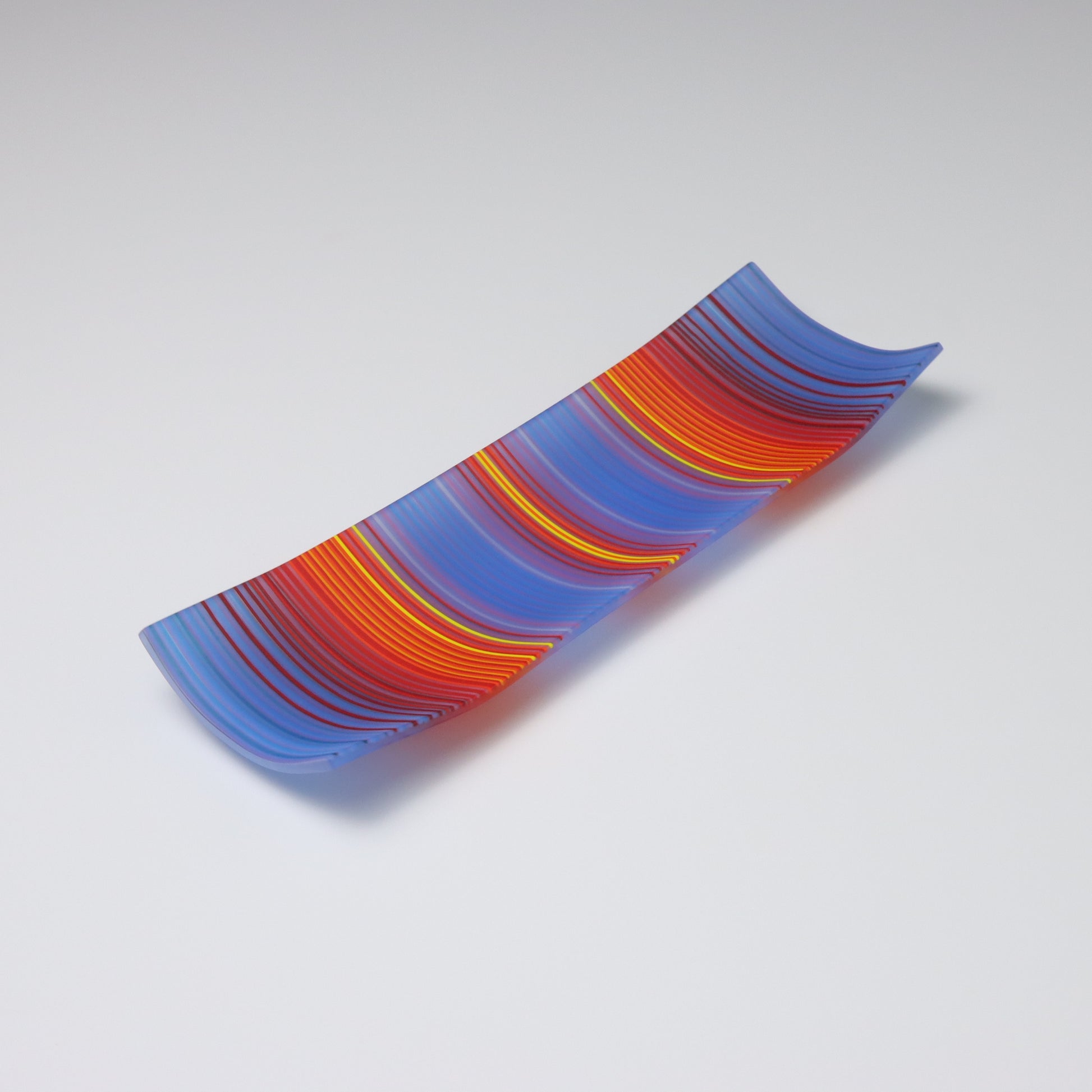 A decorative shallow rectangular boat shaped glass plate with raised corners and a  colourful ColourWave stripe patterns showing key colours of bold blue, red, orange and yellow.