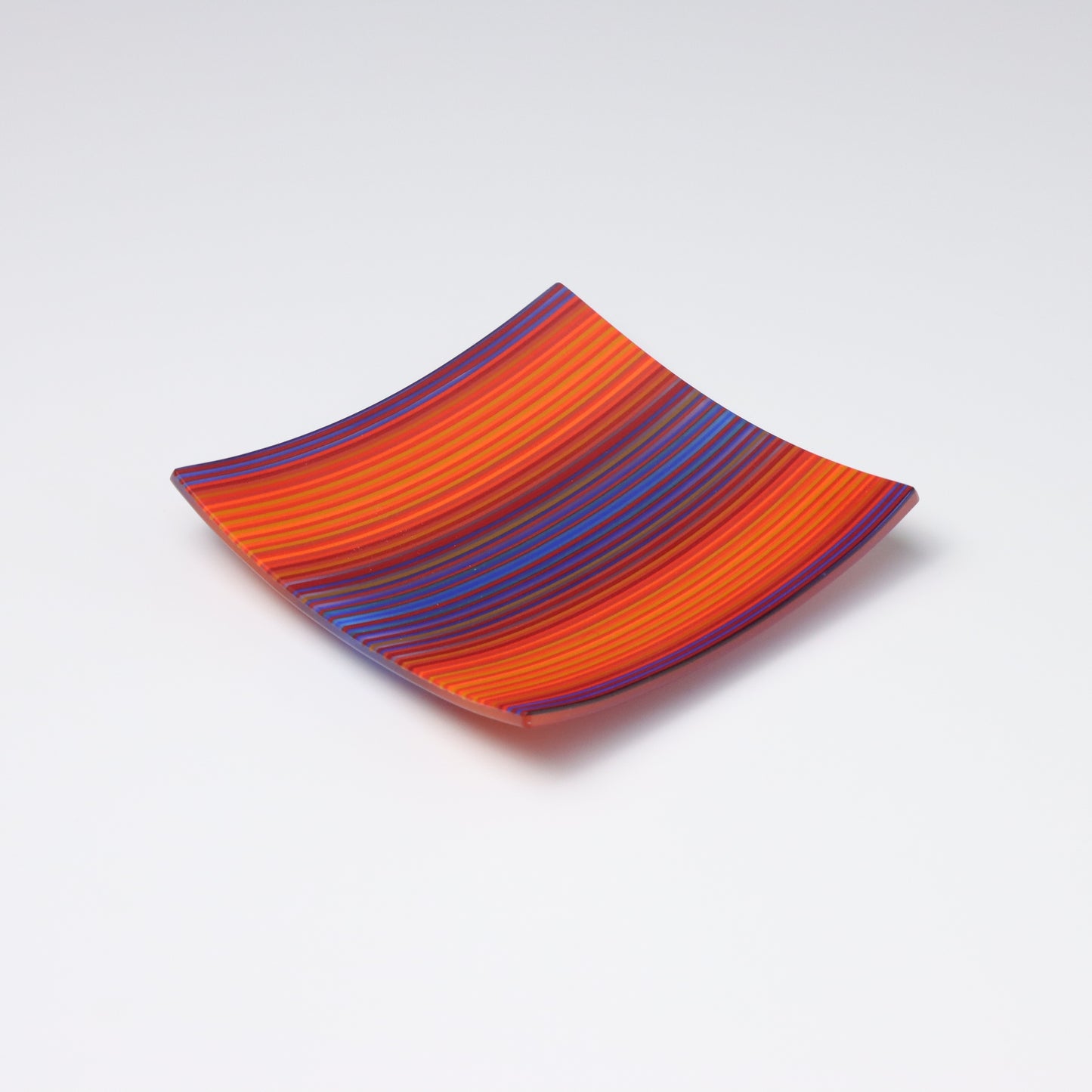 A colourful decorative fused glass small square shaped plate that curves up at the corners. With a ColourWave design the transitions from purples at the edges and center. With a warm glowing orange between.