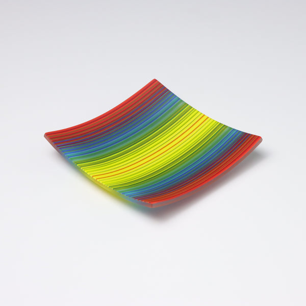 A colourful decorative fused glass small square shaped plate that curves up at the corners. With a ColourWave design that combines most of the colours of the rainbow in it, with red at the edges transitioning through blues and greens with a yellow center. 