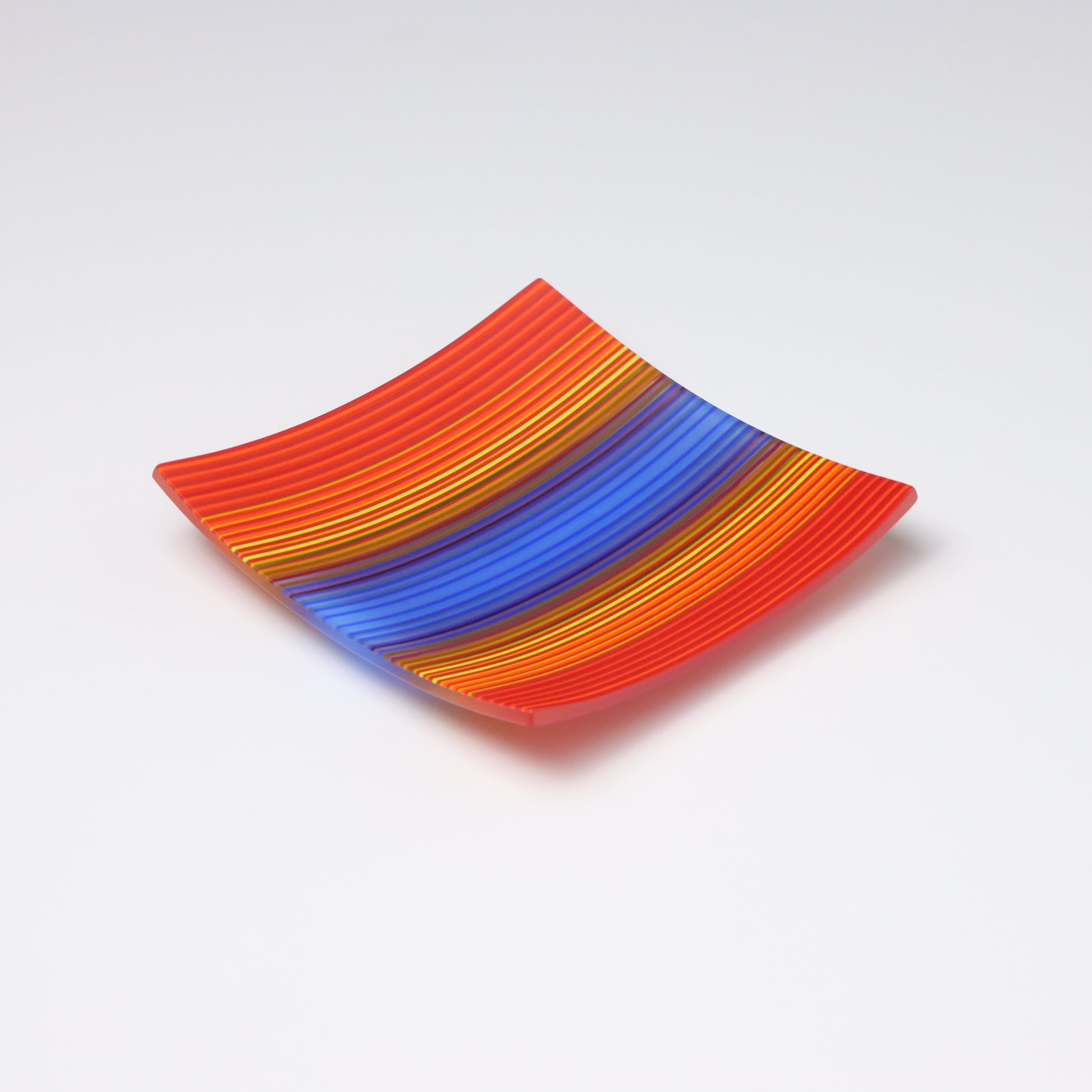 A colourful decorative glass plate - Small square shaped that curve up at the corners.  Design made up of coloured striped that start at red at the edges, transition to oranges purples and blues inthe center.